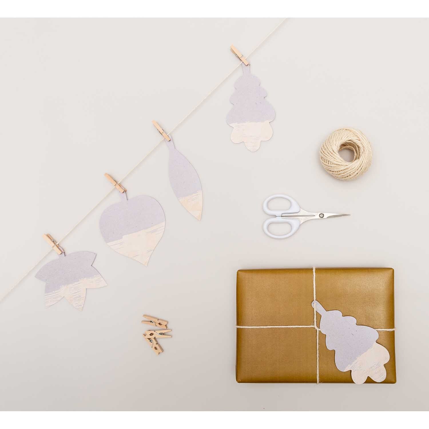 Paper Poetry Papieranhänger Blätter Graukarton, dekorativ arrangiert mit Schere, Geschenk und Kordel auf hellem Hintergrund.