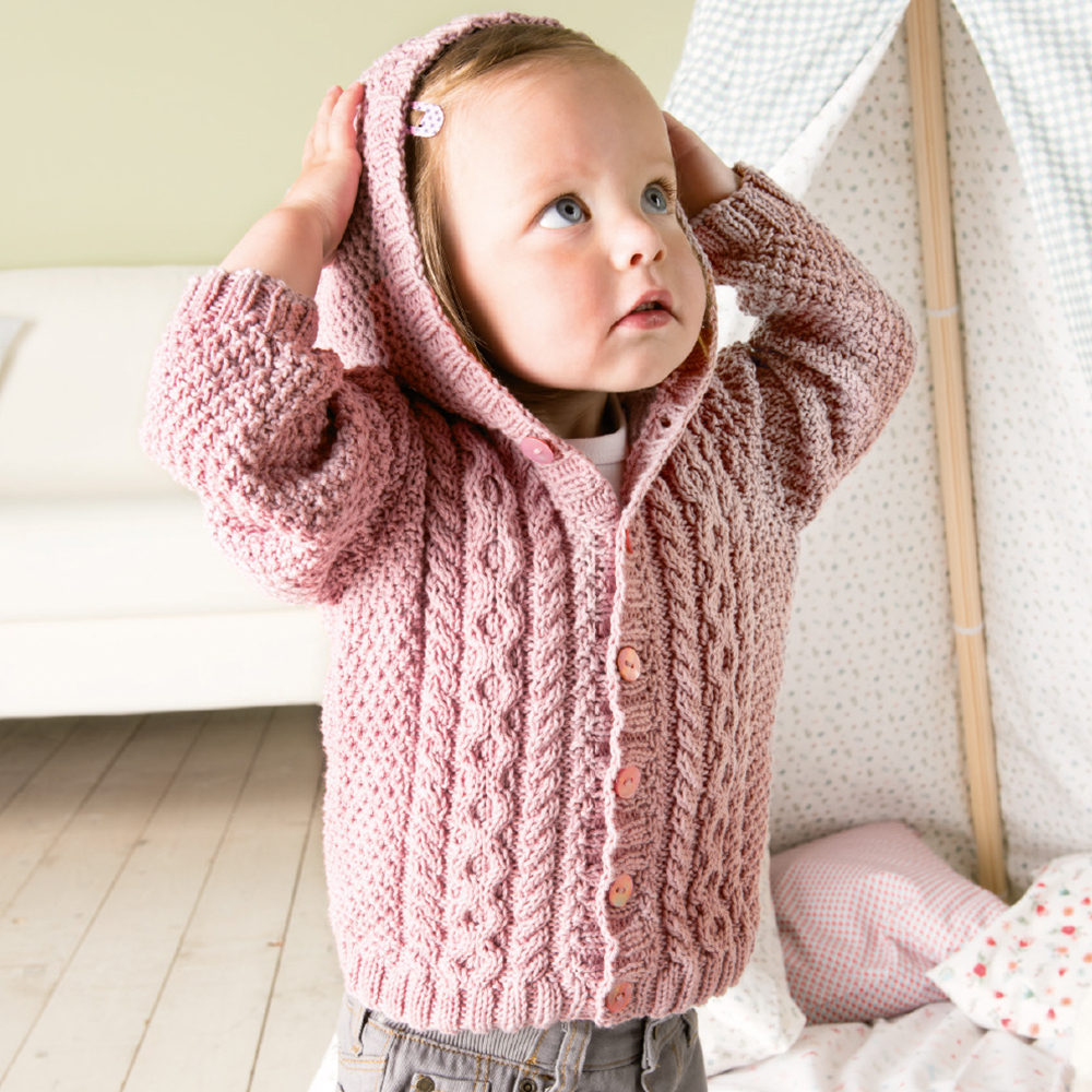 Strickanleitung Rico Baby 19 - Zopfjacke: Kleinkind in rosa Strickjacke mit Kapuze, stehend vor einem Zelt.