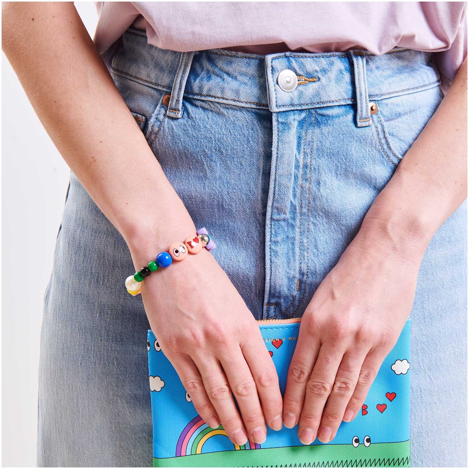 Rico Design x Redfries Armband Eye Candy Super Mix: Frau trägt buntes Perlenarmband, hält farbenfrohes Buch, trägt Jeans.