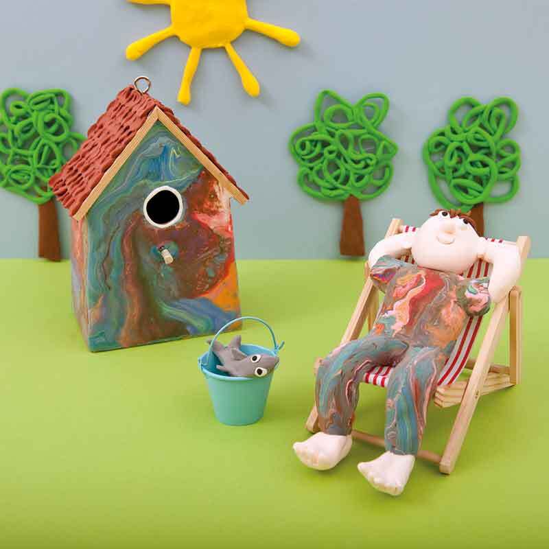 Super Fluffy 28g: Eine bunte Figur entspannt im Liegestuhl neben einem bemalten Vogelhaus.