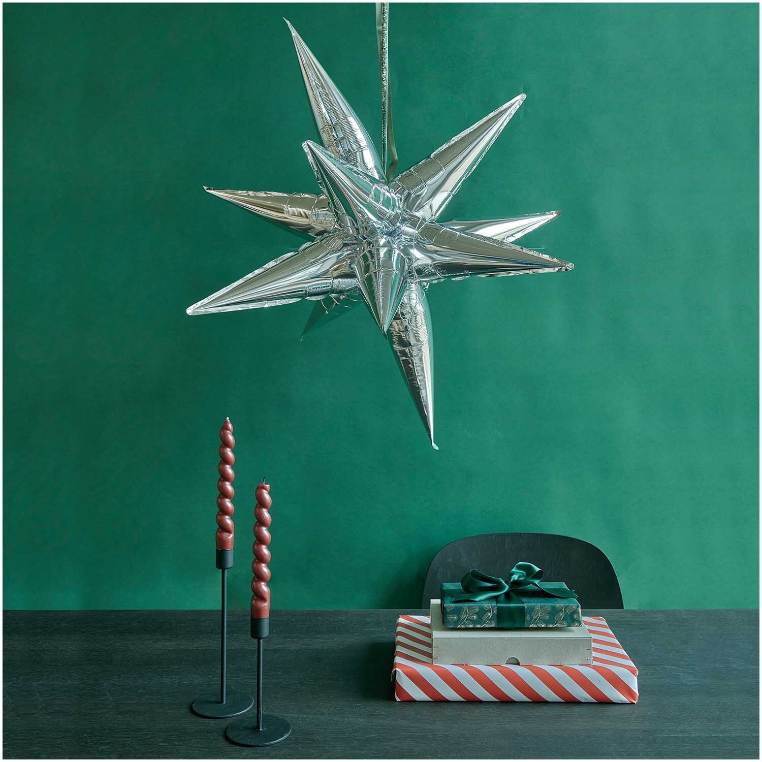 Folienballon 3D-Stern silber von Rico Design schwebt über einem Tisch mit Geschenken und Kerzen.