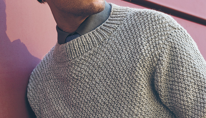 Strickanleitung Herrenpullover im Perlmuster aus Essentials Soft Merino aran. Grauer Pullover im Perlmuster, getragen von einem Mann vor einer roten Wand.