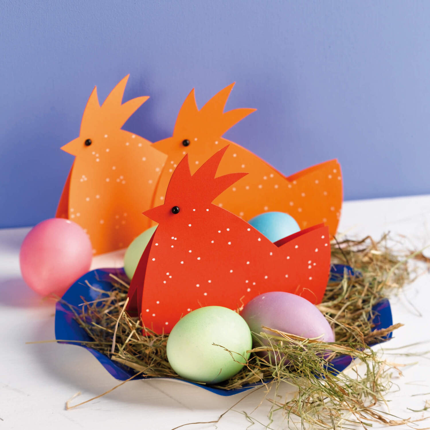 Bastelanleitung Hühner aus Papier: Drei orangefarbene Papierhühner mit bunten Eiern auf Heu.