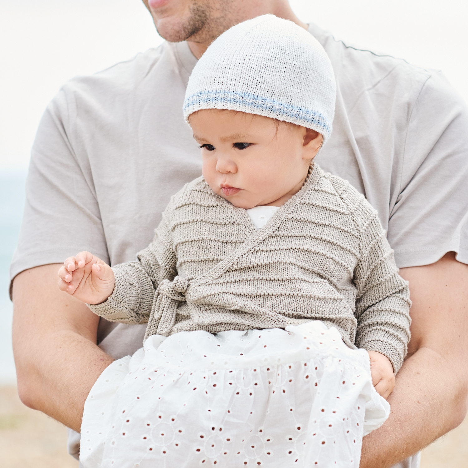 Strickanleitung Wickeljacke aus Baby Organic Cotton: Baby in grauer Wickeljacke und weißer Mütze.