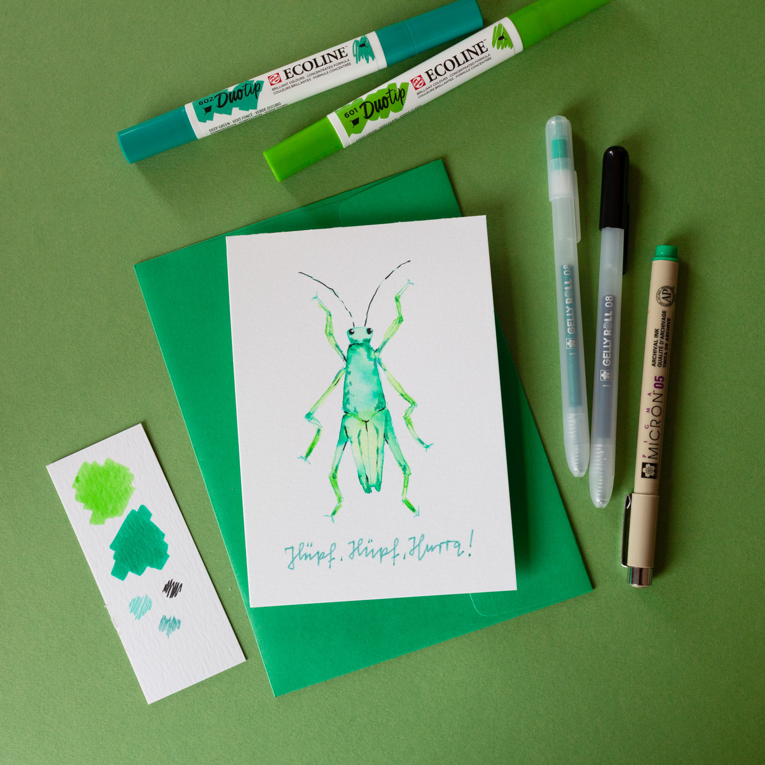 Anleitung Mixed Media Grashüpfer: Grüne Karte mit Grashüpfer-Illustration, umgeben von Stiften und Farbmuster.
