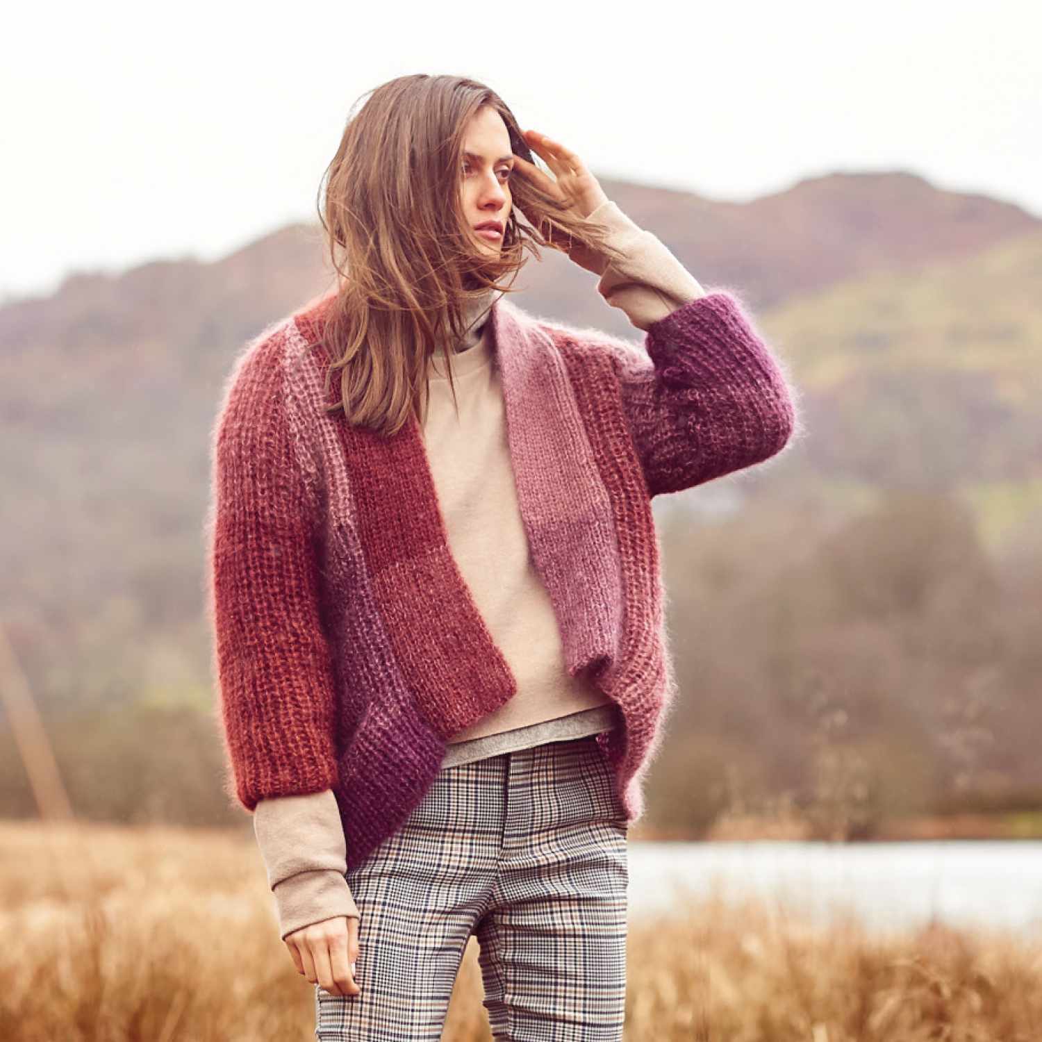 Strickanleitung Jacke aus Essentials Super Kid Mohair Loves Silk Colourlove. Frau trägt eine bunte, gestrickte Jacke in ländlicher Umgebung.