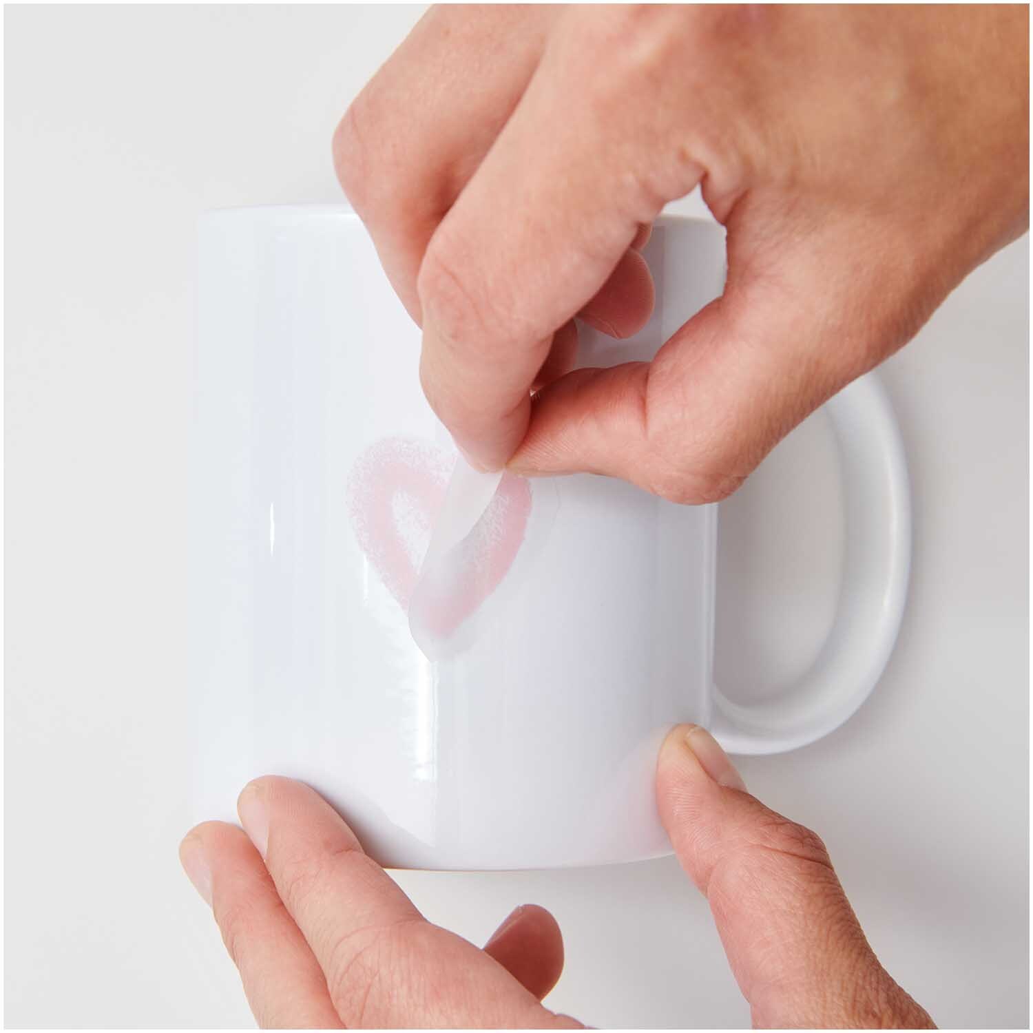 Transfermotiv Spray Herzen: Hände kleben ein Herzmotiv auf eine weiße Tasse. Marke: Rico Design.