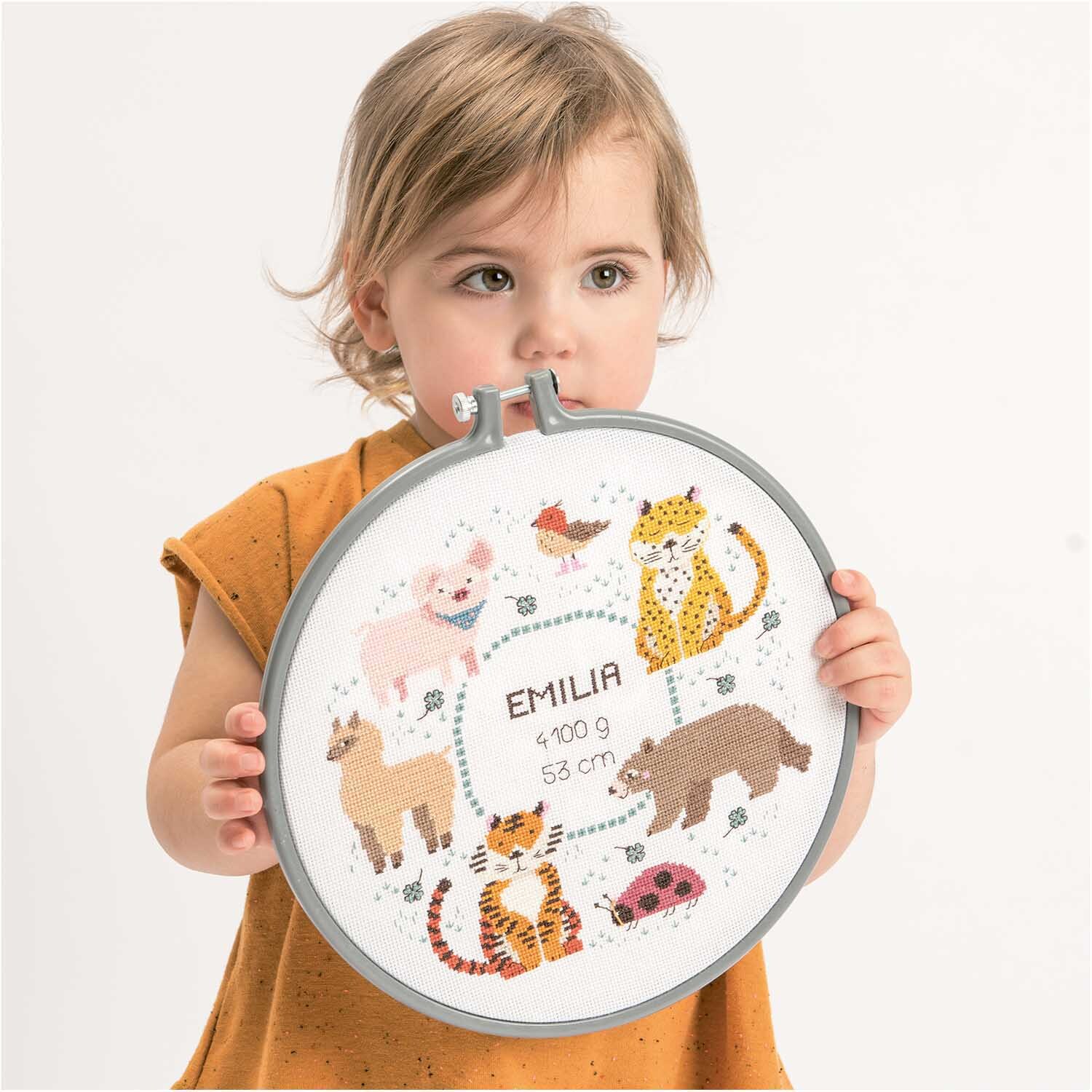 Stickpackung Geburtsbild Tiere von Rico Design zeigt ein Kind mit einem bestickten Rahmen voller Tiermotive.