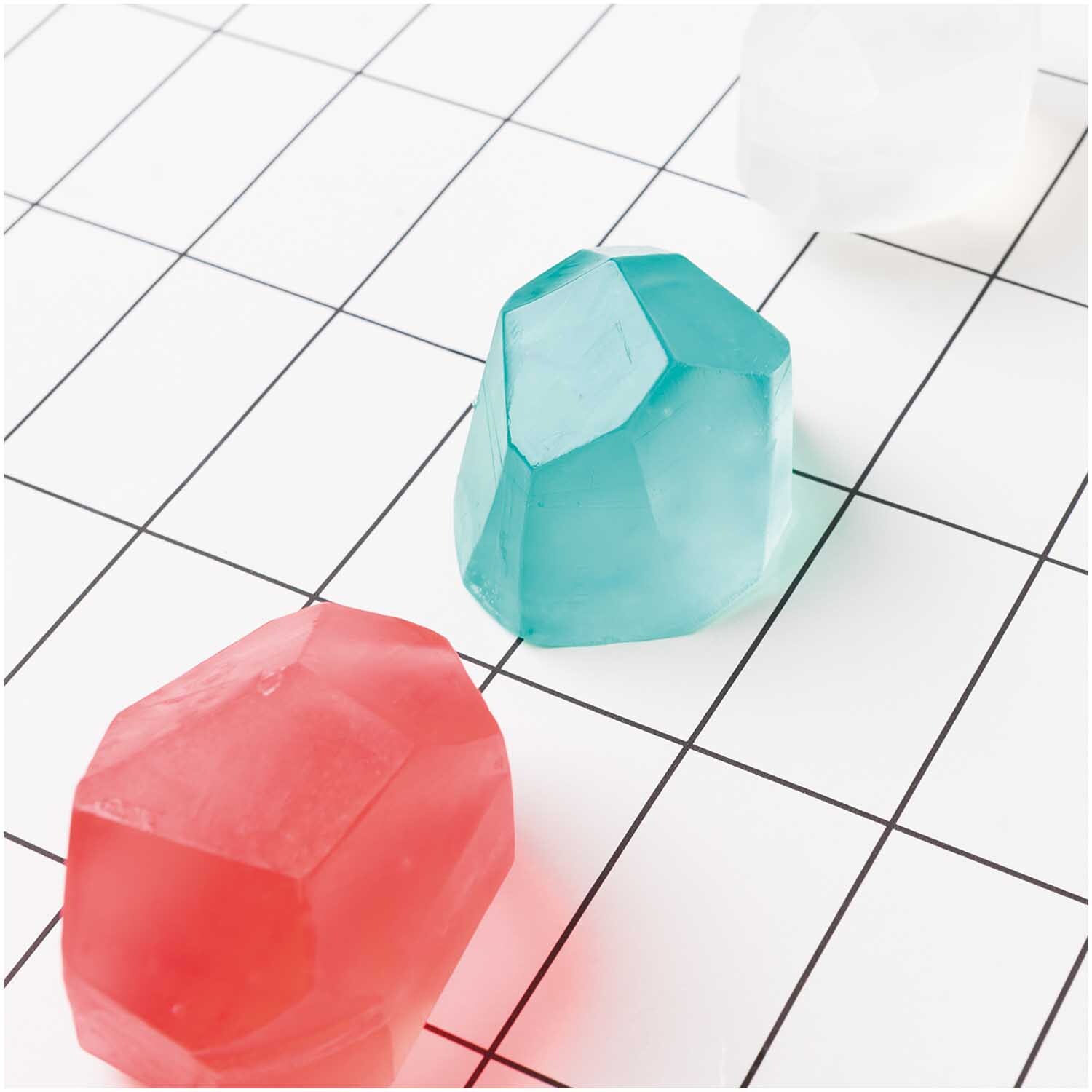 Gießseife transparent in Edelsteinform auf kariertem Hintergrund. Farben: Rot, Blau, Weiß.