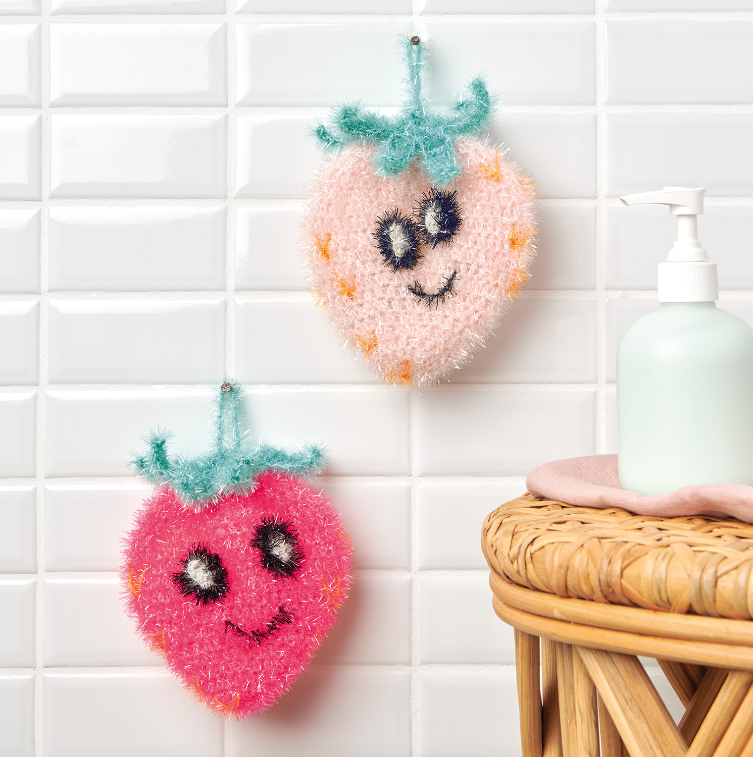 Häkelanleitung Erdbeere aus Creative Bubble: Zwei gehäkelte Erdbeeren mit Gesichtern hängen an einer weißen Fliesenwand.