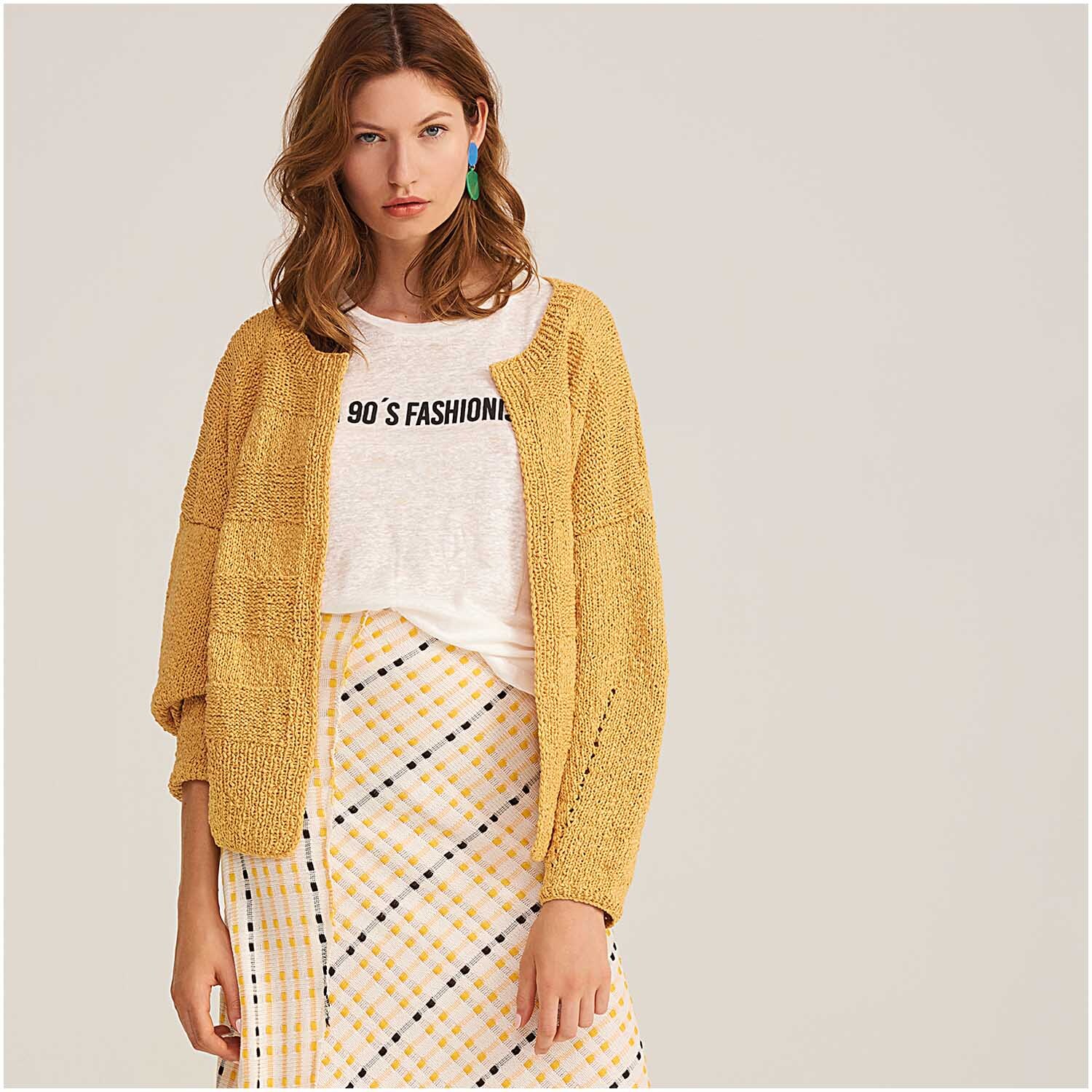 Fashion Linen Swell aran: Frau in senffarbener Strickjacke und kariertem Rock, Marke Rico Design.