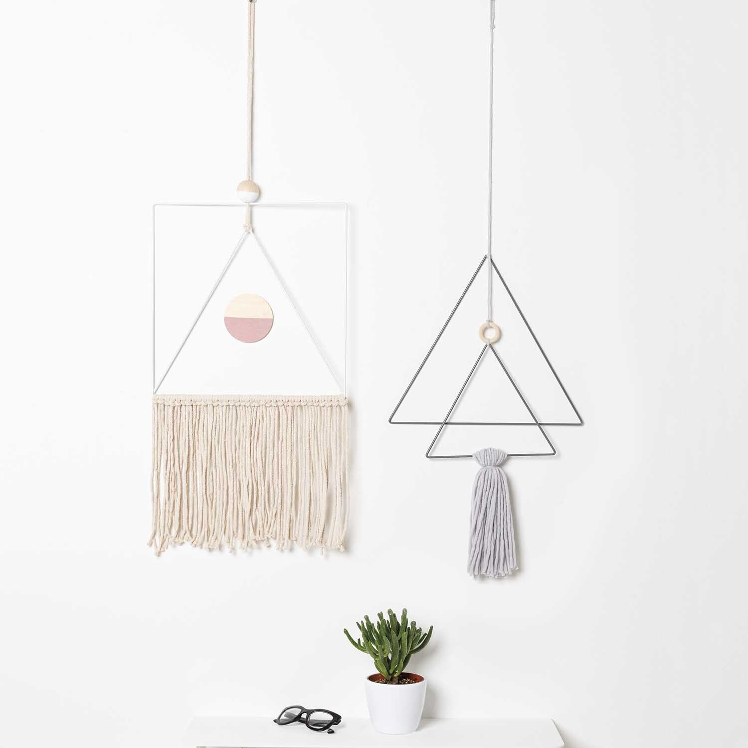 DIY Set Makramee: Zwei geometrische Makramee-Wandbehänge von Rico Design, einer mit Fransen, der andere mit Quaste, neben einer Pflanze.