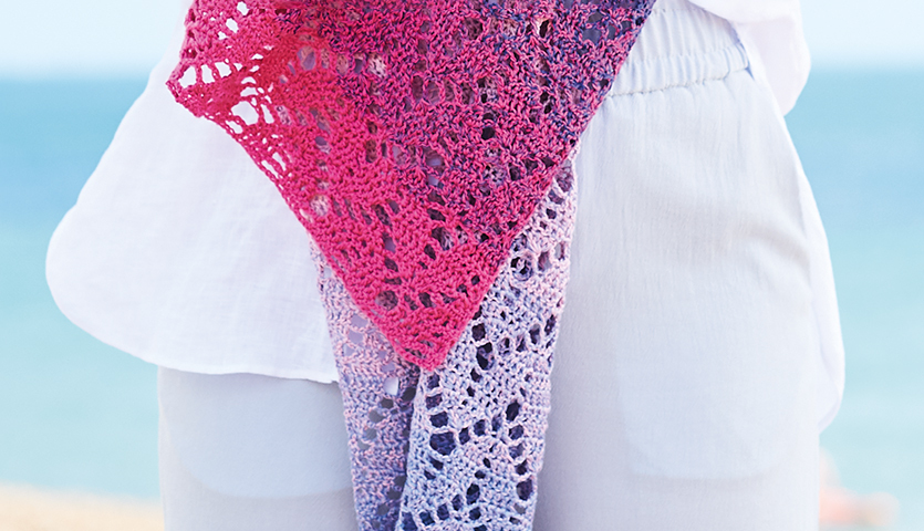 Häkelanleitung Tuch aus Creative Cotton Dégradé Lucky 8: Frau trägt ein gehäkeltes Tuch in Rosa- und Lilatönen am Strand.