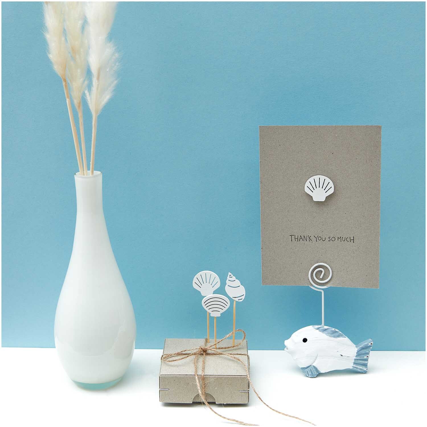Deko-Picker Muschel Mix in elegantem Weiß von Rico Design, arrangiert neben einer Vase und Geschenkbox.