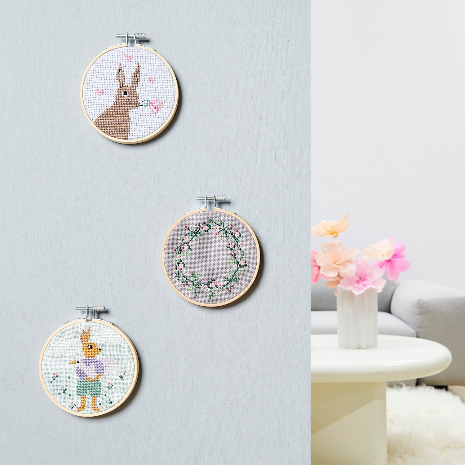 Stickpackung Bild Hase mit Gans im Arm: Drei Stickrahmen an der Wand, darunter ein Hase mit Gans und ein Blumenkranz. Marke: Rico Design.