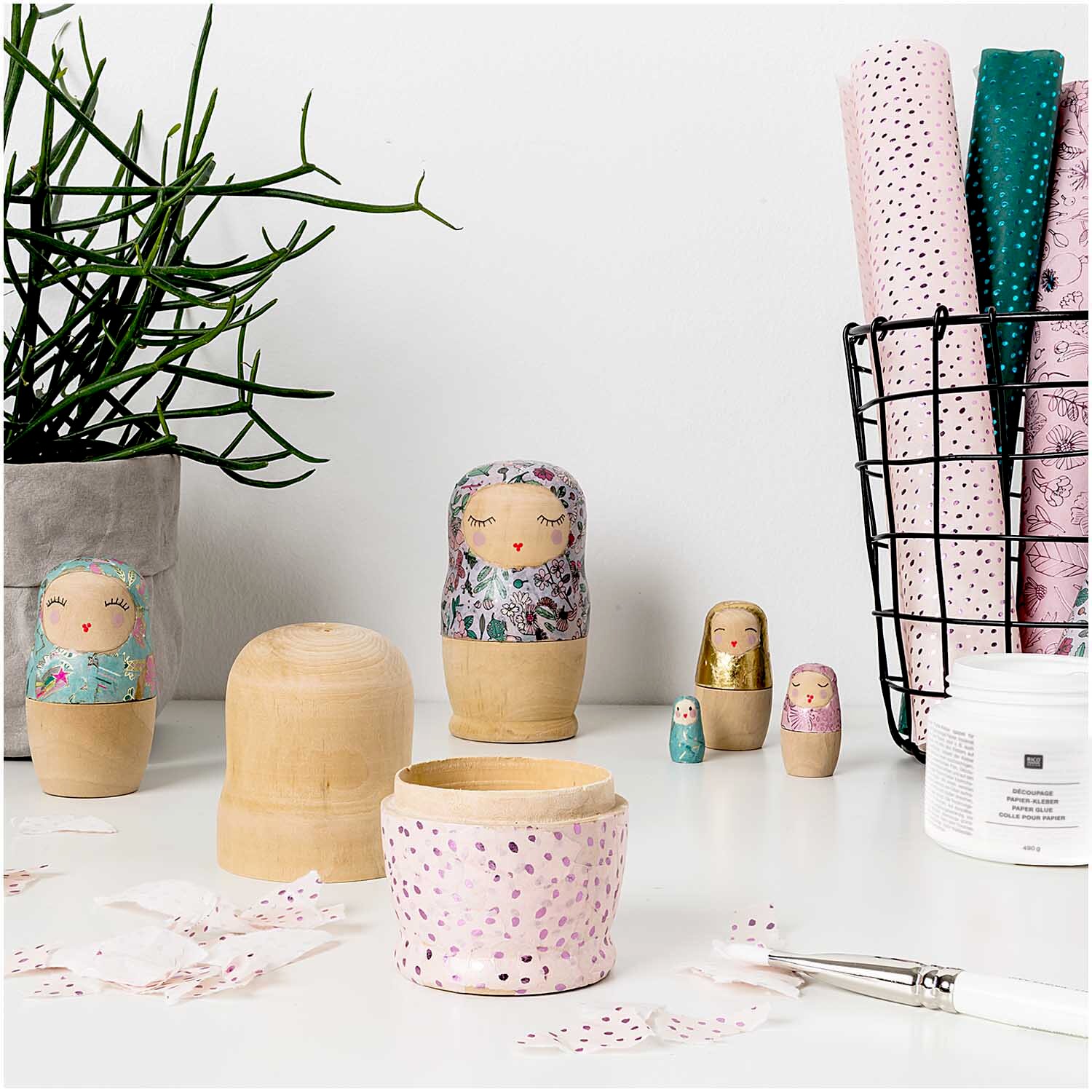 Paper Poetry Kleber von Rico Design auf Basteltisch mit Holz-Matrjoschkas, Pinsel, Stoffresten und dekorativen Papieren für kreative DIY-Projekte