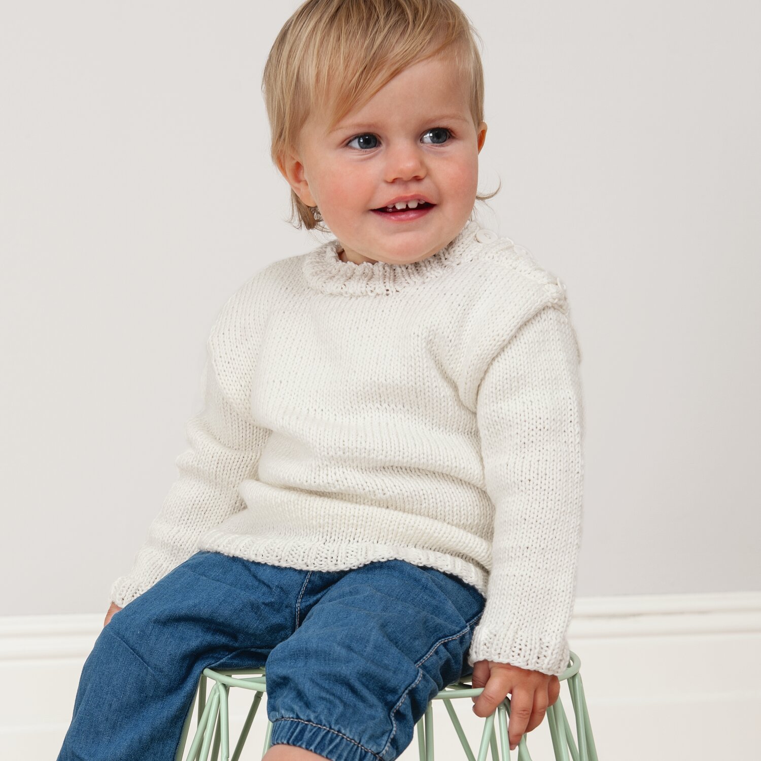 Strickset Babypullover weiß von Rico Design: Ein Baby trägt einen gestrickten Pullover, sitzt auf einem Stuhl und lächelt.
