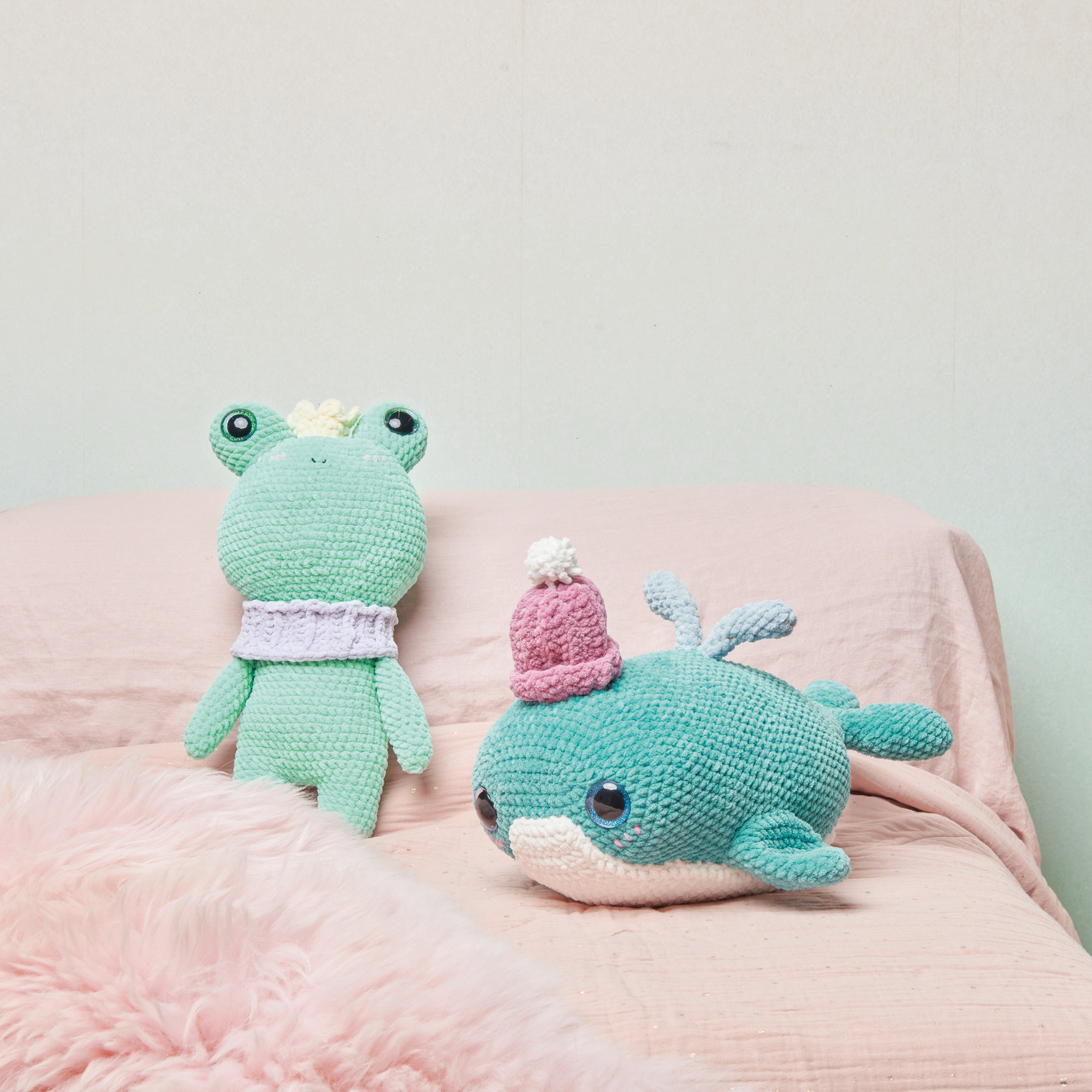 Häkelset Cute Crowned Frog: Ein grüner Frosch mit Krone sitzt neben einem gehäkelten Wal auf einem rosa Bett.