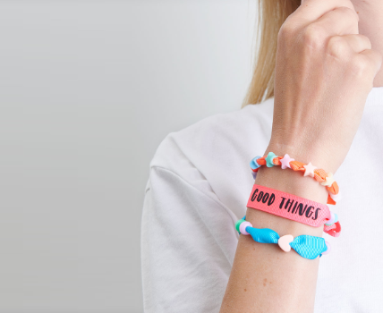 Armgelenk mit einem orangen Armband mit bunten Sternen, ein pinkes Armband mit schwarzem Schriftzug und einem blauen Armband mit Herz und runder Perle.