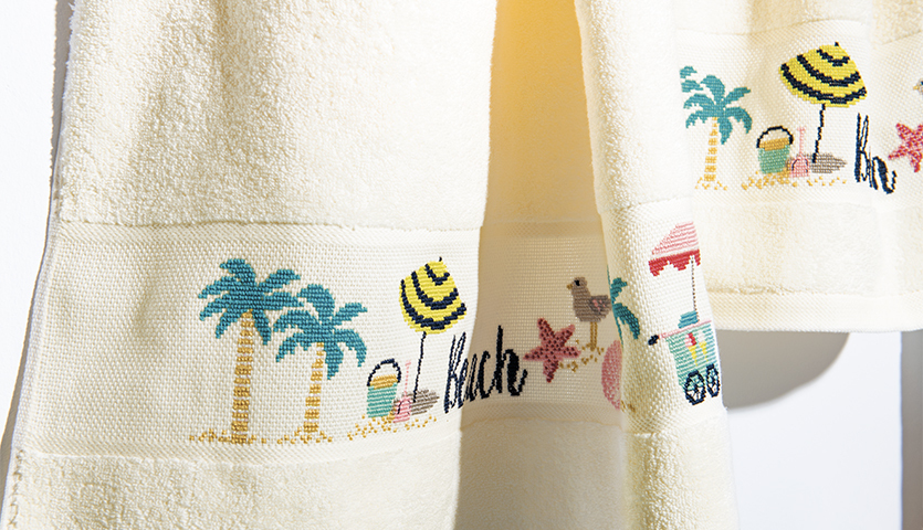 Stickanleitung Frottierhandtuch mit Strand-Motiven, zeigt Palmen, Sonnenschirme und Strandspielzeug auf hellem Stoff.