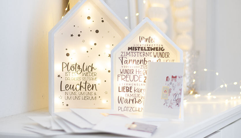 Inspiration Achtsamkeits-Adventskalender: Zwei beleuchtete Deko-Häuser mit Schriftzügen auf weißem Regal, umgeben von Lichterketten.