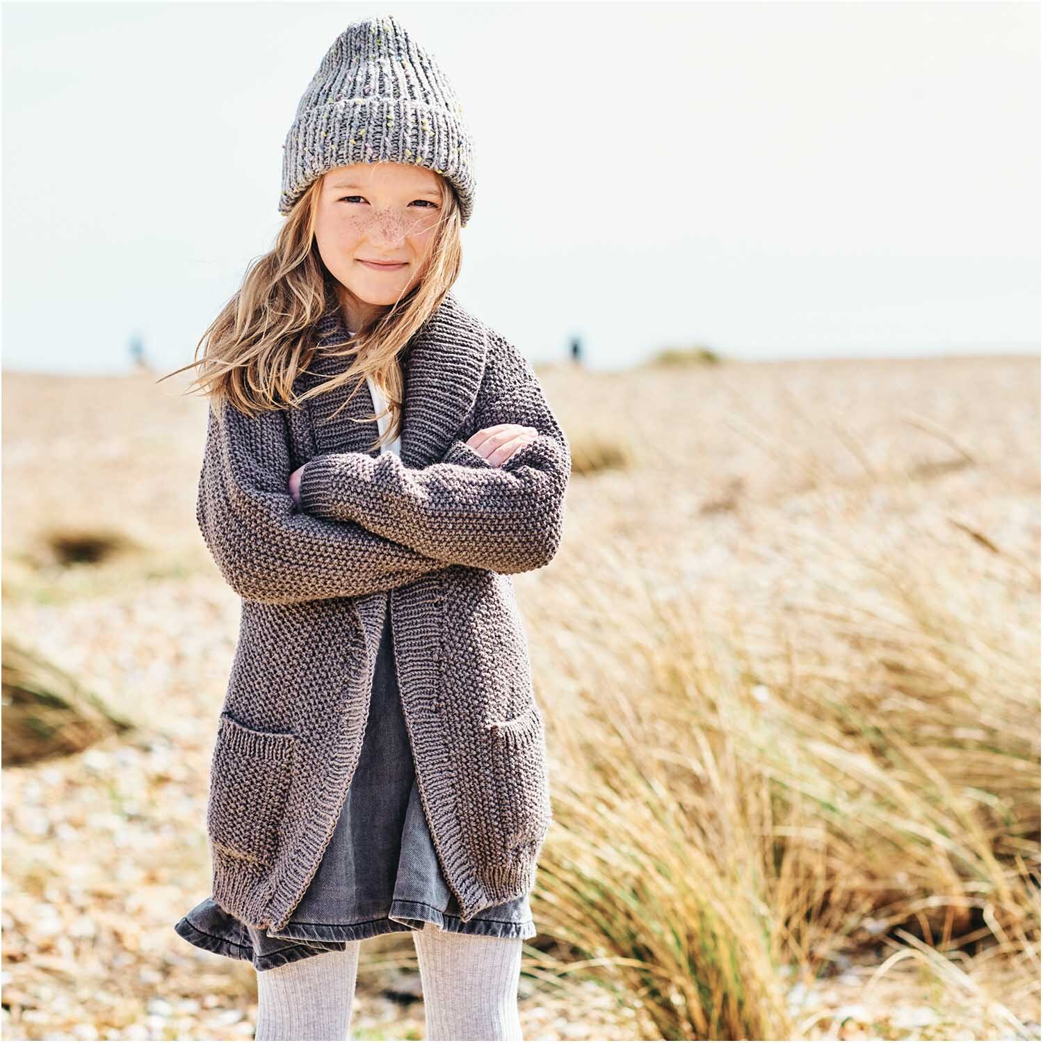 Creative Jazz Up: Mädchen in gestricktem Outfit am Strand, trägt graue Mütze und Strickjacke.