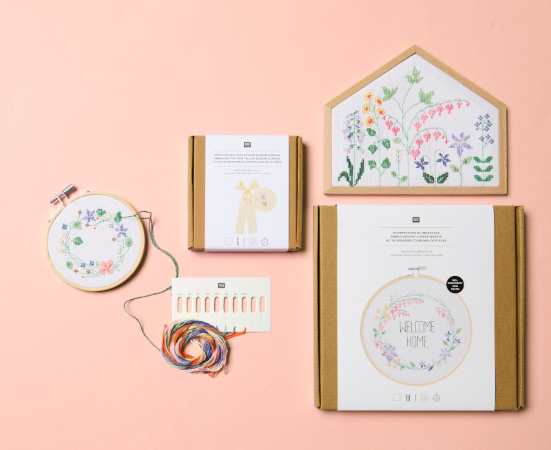 Eine Stickpackung mit Stickring und Blumenkranz Motiv, sowie ein Stickrahmen in Hausform mit Blumenwiese Motiv. Flatlay von Stickpackungen.