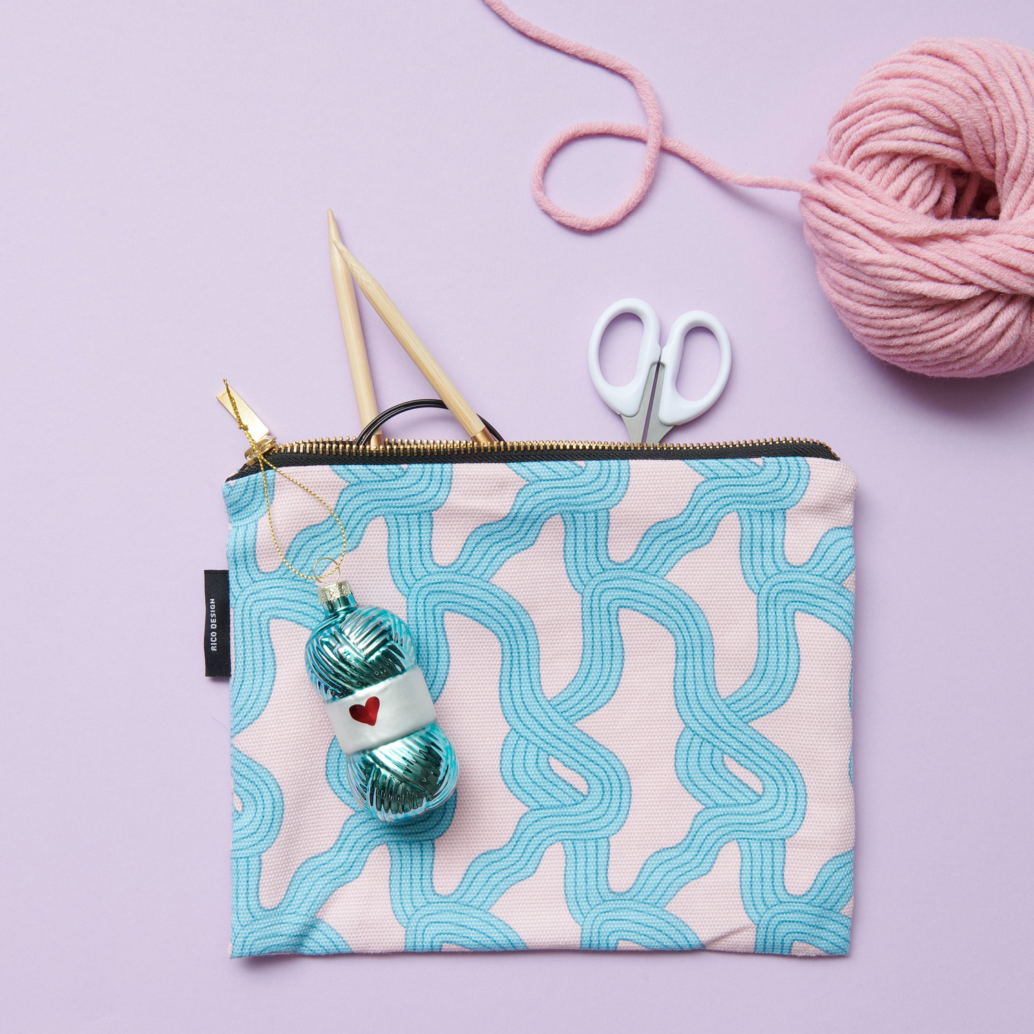 Canvas-Pouches Myzel Rosa mit blauem Muster, gefüllt mit Stricknadeln, Schere und Garn auf lila Hintergrund. Größe: 17x23cm.