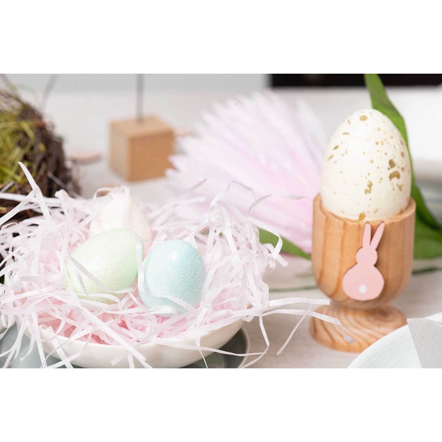 DIY Set Osterdeko pastell: Pastellfarbene Ostereier in einem Nest aus Papierstreifen, daneben ein Holz-Eierbecher mit Hasenmotiv.