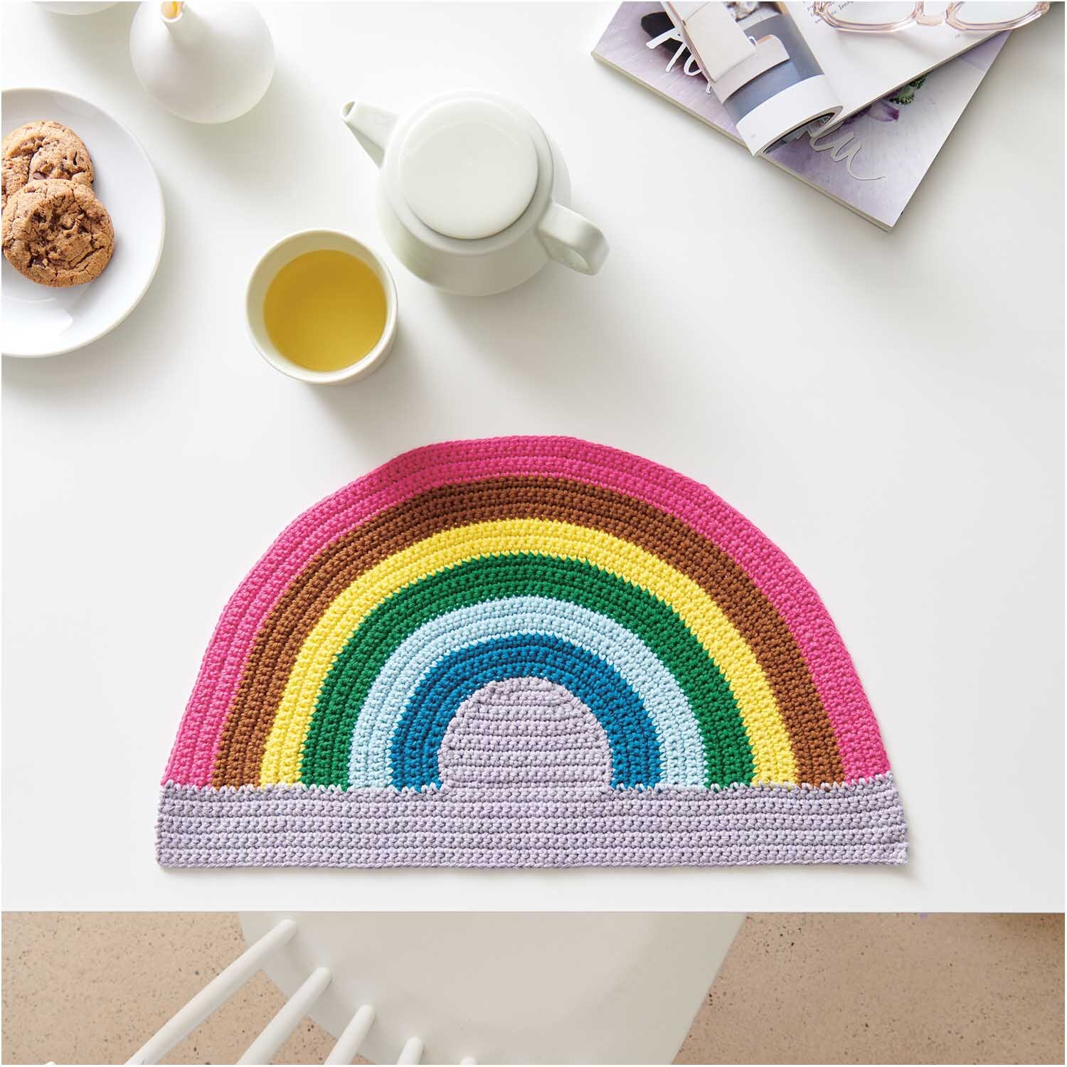 Cotton Universal: Gehäkelter Regenbogen-Topflappen auf weißem Tisch mit Tee und Keksen, Wolle Rödel.