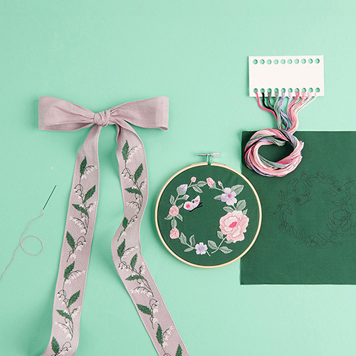 Eine Schleife mit Maiglöckchen bestickt und ein Stickring mit einem Blumenkranz bestickt, daneben passendes Zubehör.
