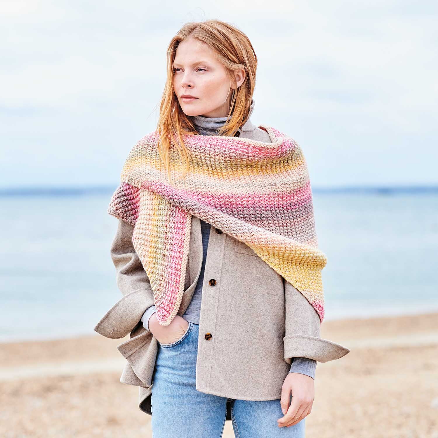 Strickanleitung Dreieckstuch aus Chic-Unique: Frau am Strand mit pastellfarbenem Tuch über beigem Mantel.
