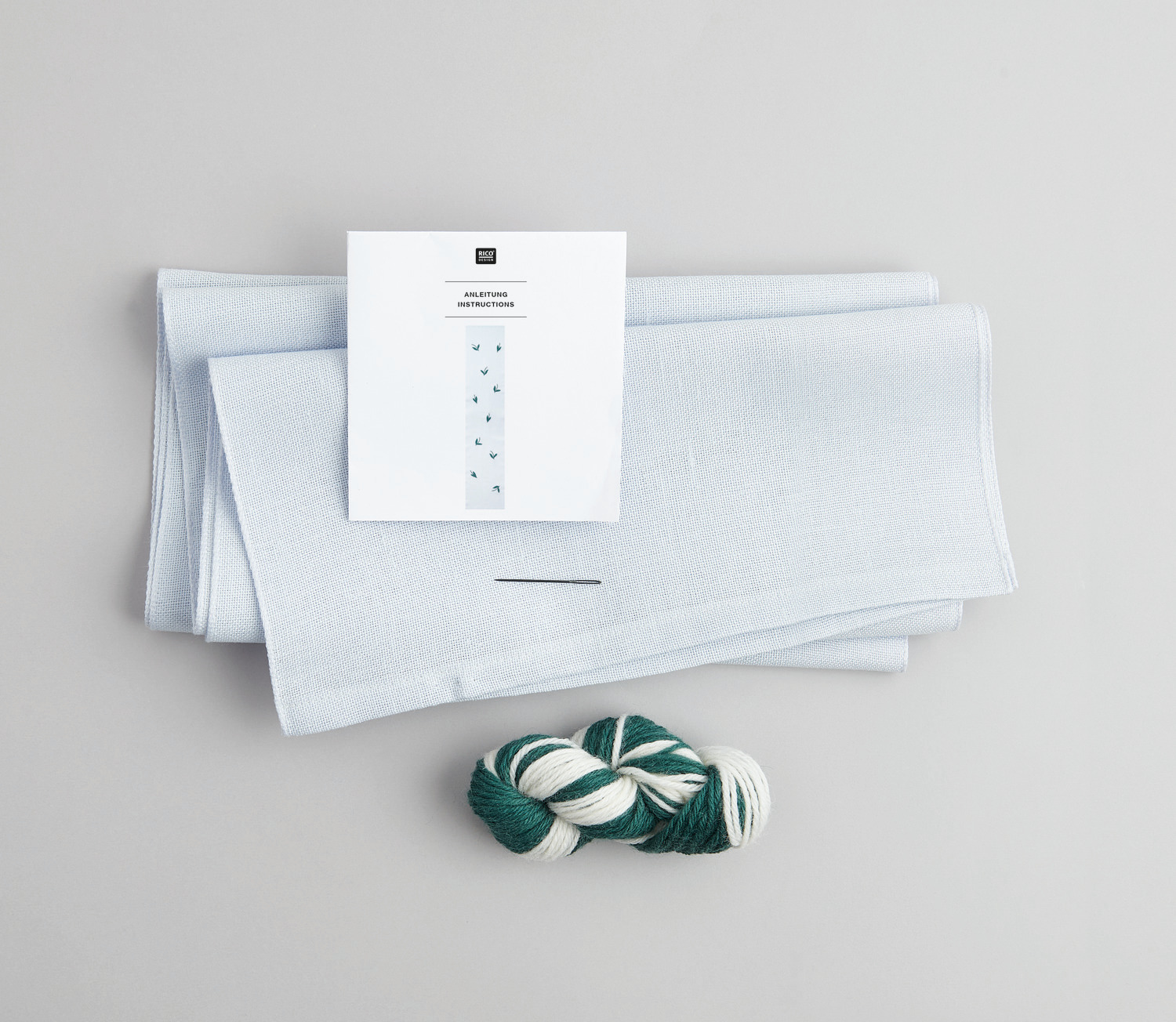 Stickpackung Tischband Maiglöckchen vorgezeichnet von Rico Design, mit Garn und Nadel auf grauem Hintergrund.