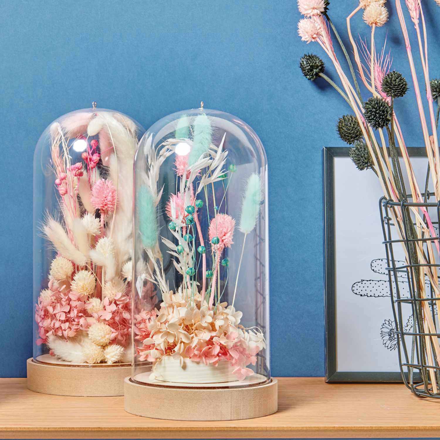 Bastelanleitung Dekohauben mit Trockenblumen: Zwei Glasglocken mit bunten Trockenblumen auf Holzsockeln vor blauer Wand.