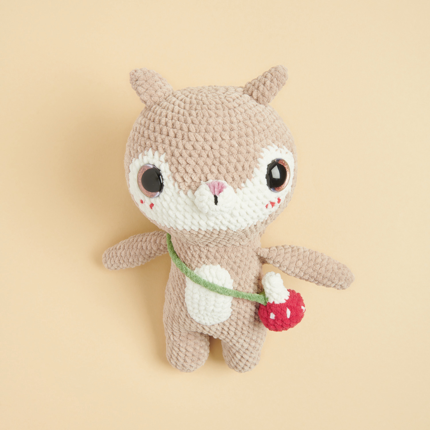 Häkelset Little Squirrel Modell 02: Beiges Plüschtier-Eichhörnchen mit roter Tasche auf hellem Hintergrund.
