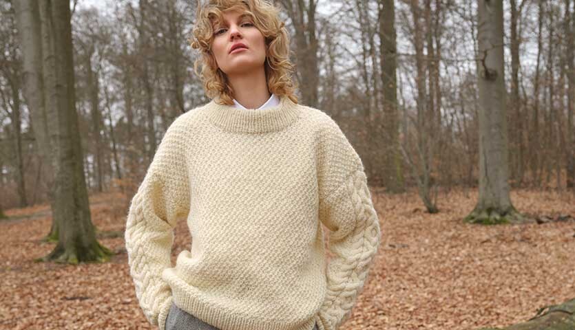 Strickanleitung Pullover aus Essentials Organic Wool aran: Frau im cremefarbenen Pullover im herbstlichen Wald.