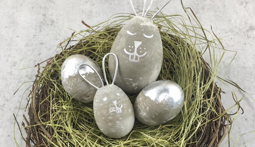 Bastelanleitung Osterhasen und Ostereier aus Beton in einem Nest aus Stroh auf grauem Hintergrund.