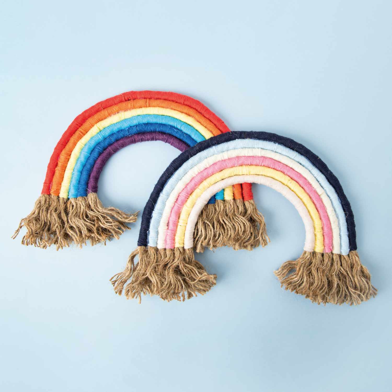 Bastelanleitung Woll-Regenbogen: Zwei handgefertigte Regenbögen aus bunter Wolle mit Fransen auf hellblauem Hintergrund.