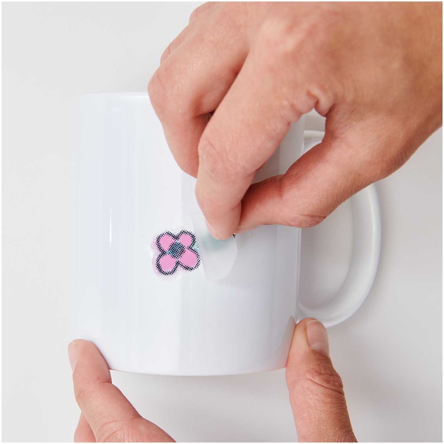 Transfermotiv Spray-Mix 10 Stück: Hände kleben ein buntes Blumenmotiv auf eine weiße Tasse. Marke: Rico Design.