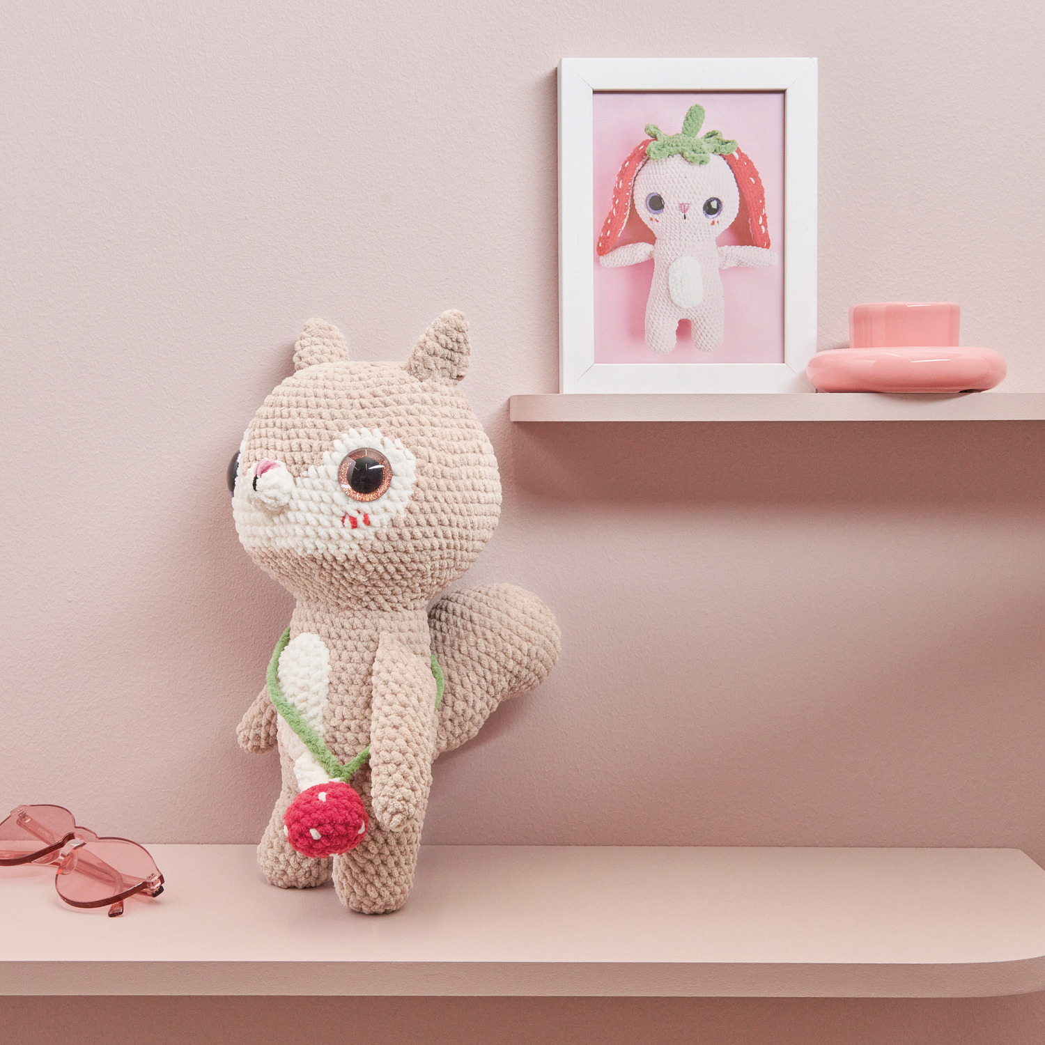 Häkelset Little Squirrel Modell 02: Beiges gehäkeltes Eichhörnchen mit roter Tasche vor rosa Wand.