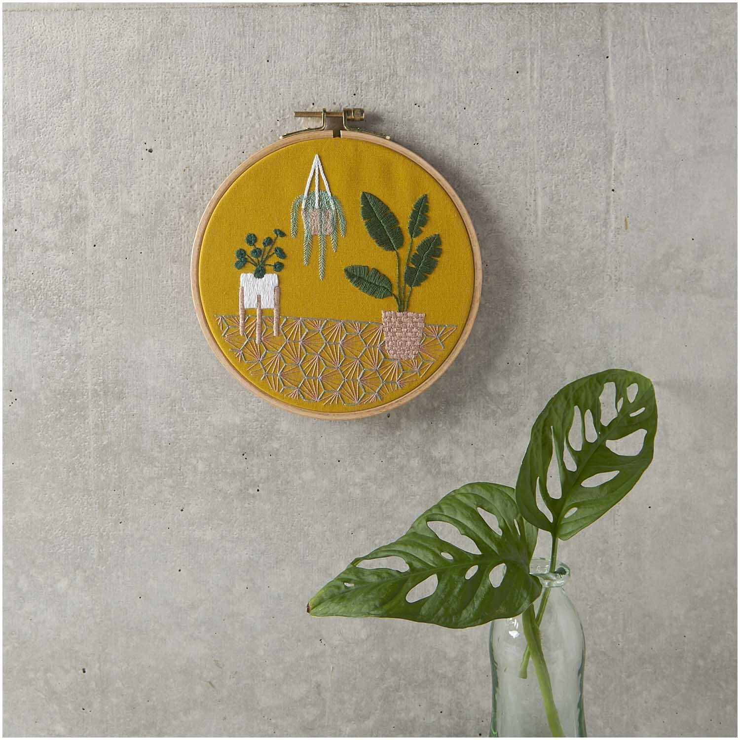Stickpackung Slow Evenings Urban Jungle Bild Ø 15,5cm zeigt Pflanzenstickerei auf gelbem Stoff, aufgehängt an grauer Wand.