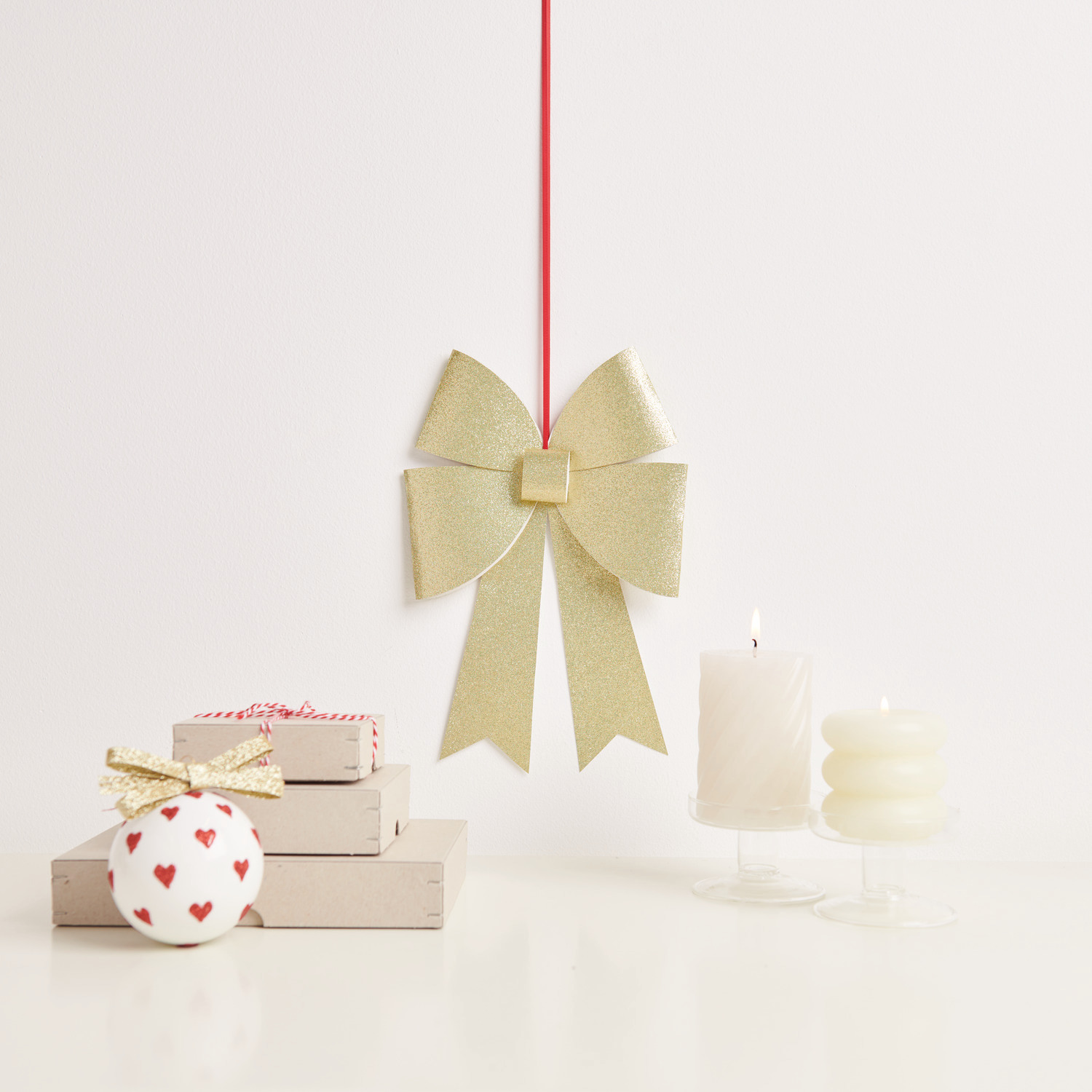 Paper Poetry Bastelset XL-Schleife Glitter: Goldene Schleife von Rico Design, dekorativ an rotem Band hängend, neben Geschenken und Kerzen.