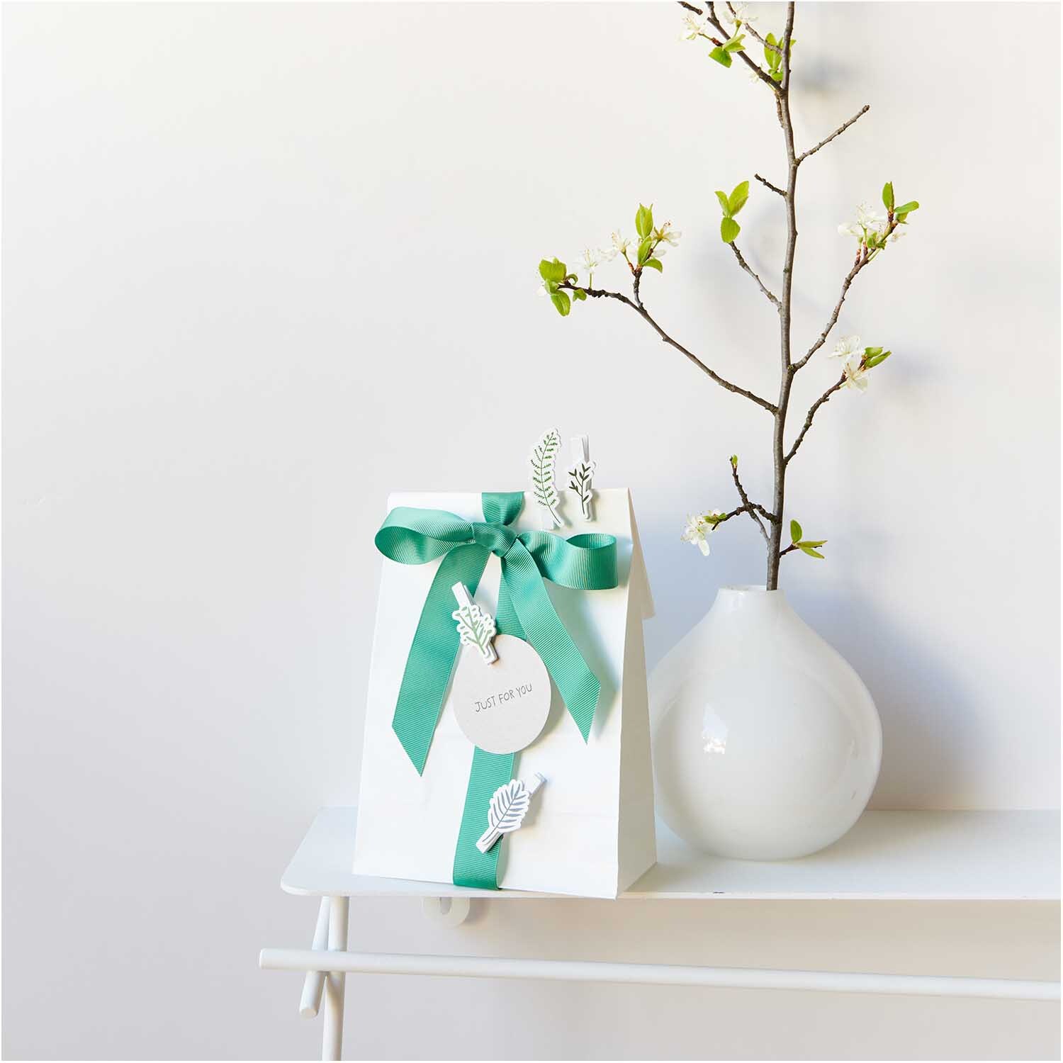 Dekoklammern Blätter Mix 4 Stück: Geschenkverpackung mit grüner Schleife und Blattklammern, dekoriert auf einem weißen Tisch neben einer Vase.