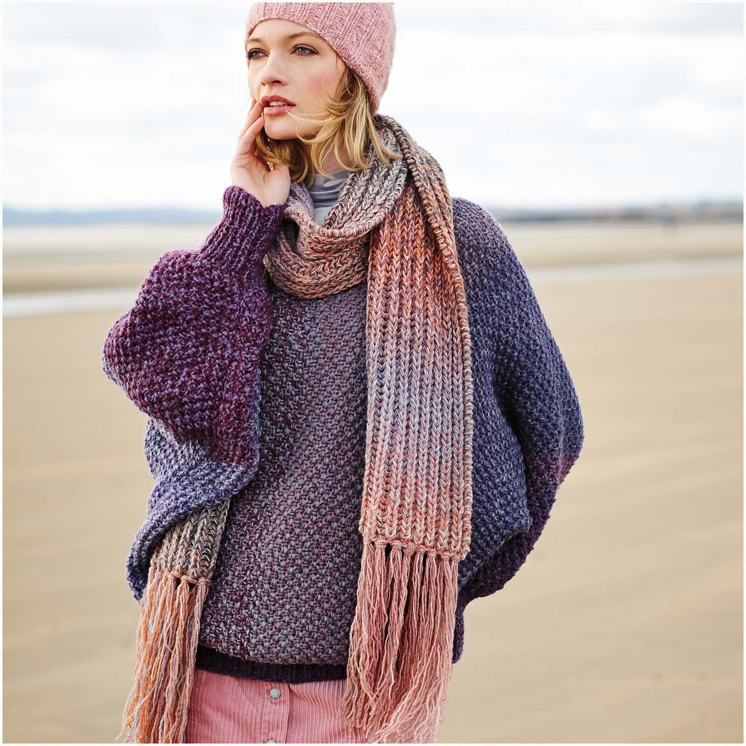 Creative Wool dégradé: Frau am Strand in gestricktem Schal und Mütze in Rosa- und Lilatönen.