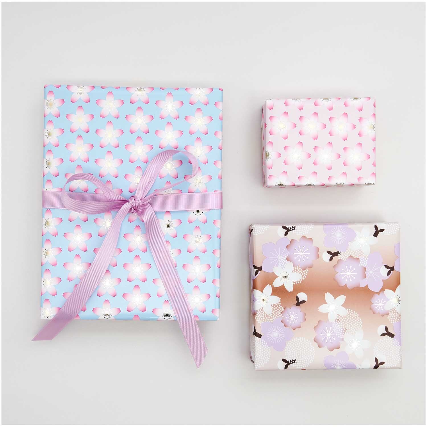 Geschenkpapier Kirschblüten Pink: Drei Geschenke in pastellfarbenem Papier mit floralen Mustern, verziert mit einer rosa Schleife.