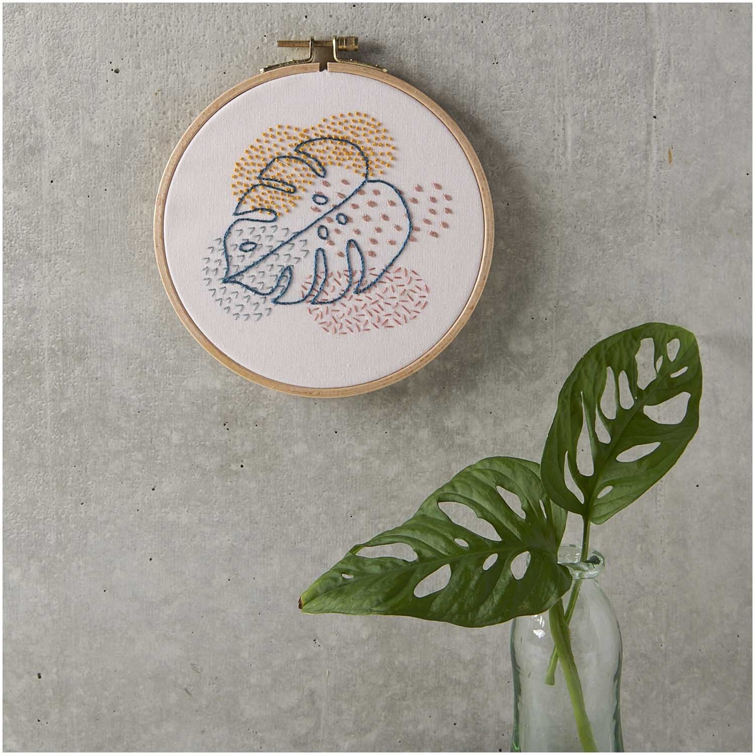 Stickpackung Slow Evenings Monstera Bild Ø 15,5cm zeigt ein gesticktes Monstera-Motiv in bunten Farben an einer grauen Wand.