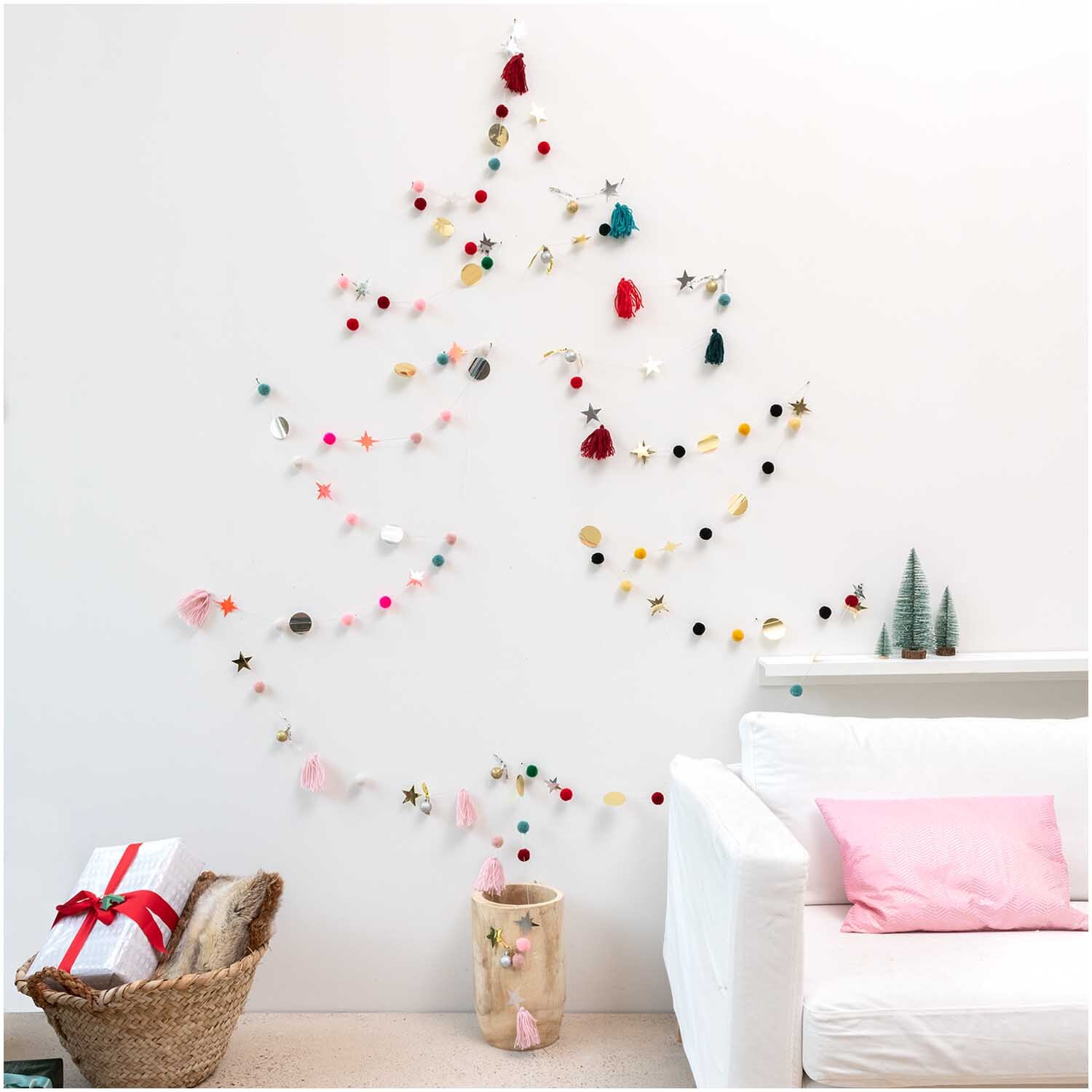 Quasten-Girlande rosa-pastell 2,2m dekoriert eine Wand in Form eines Weihnachtsbaums, umgeben von Geschenken und einem Sofa.