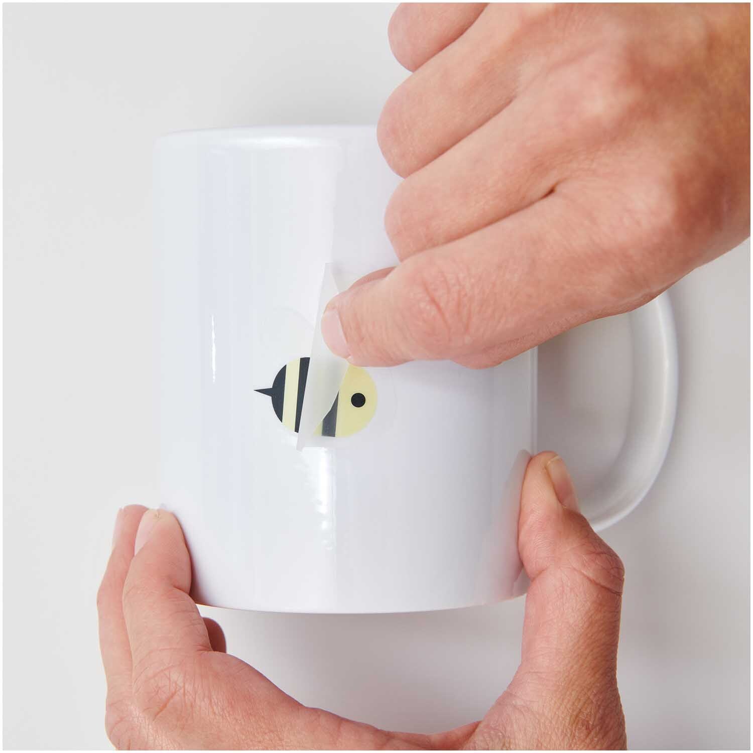 Transfermotiv Biene: Hände kleben ein Bienenmotiv von Rico Design auf eine weiße Tasse.