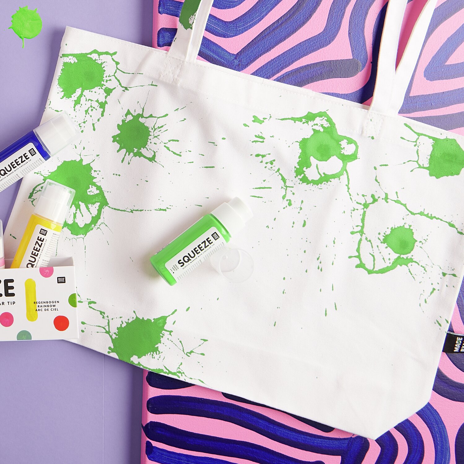 Squeeze Acrylic Marker 60ml: Stofftasche mit grünen Farbspritzern, umgeben von bunten Acrylmarkern. Ideal für kreative Designs.