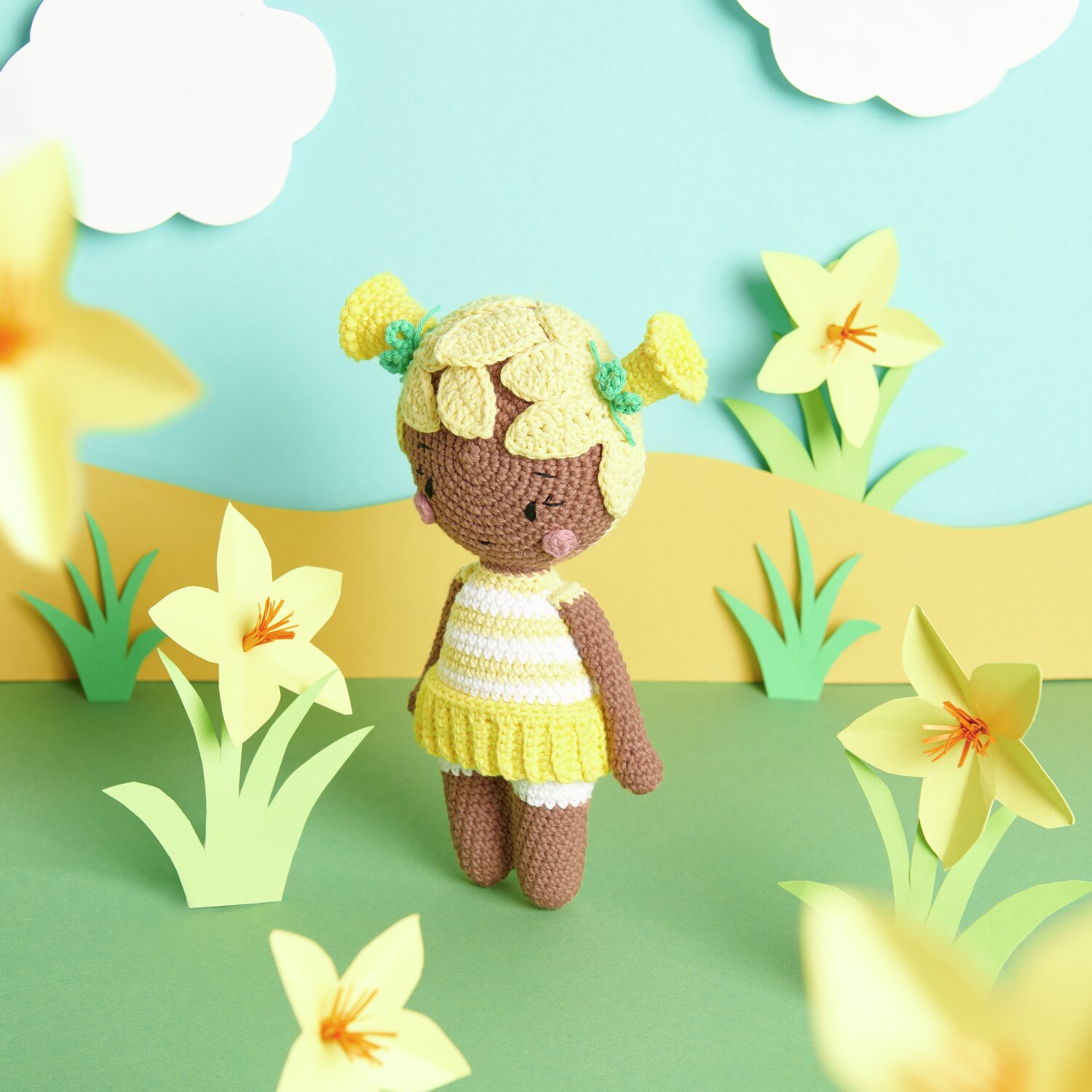 Creative Ricorumi dk: Gehäkelte Puppe in gelb-weißem Outfit, umgeben von gelben Blumen auf grünem Hintergrund.